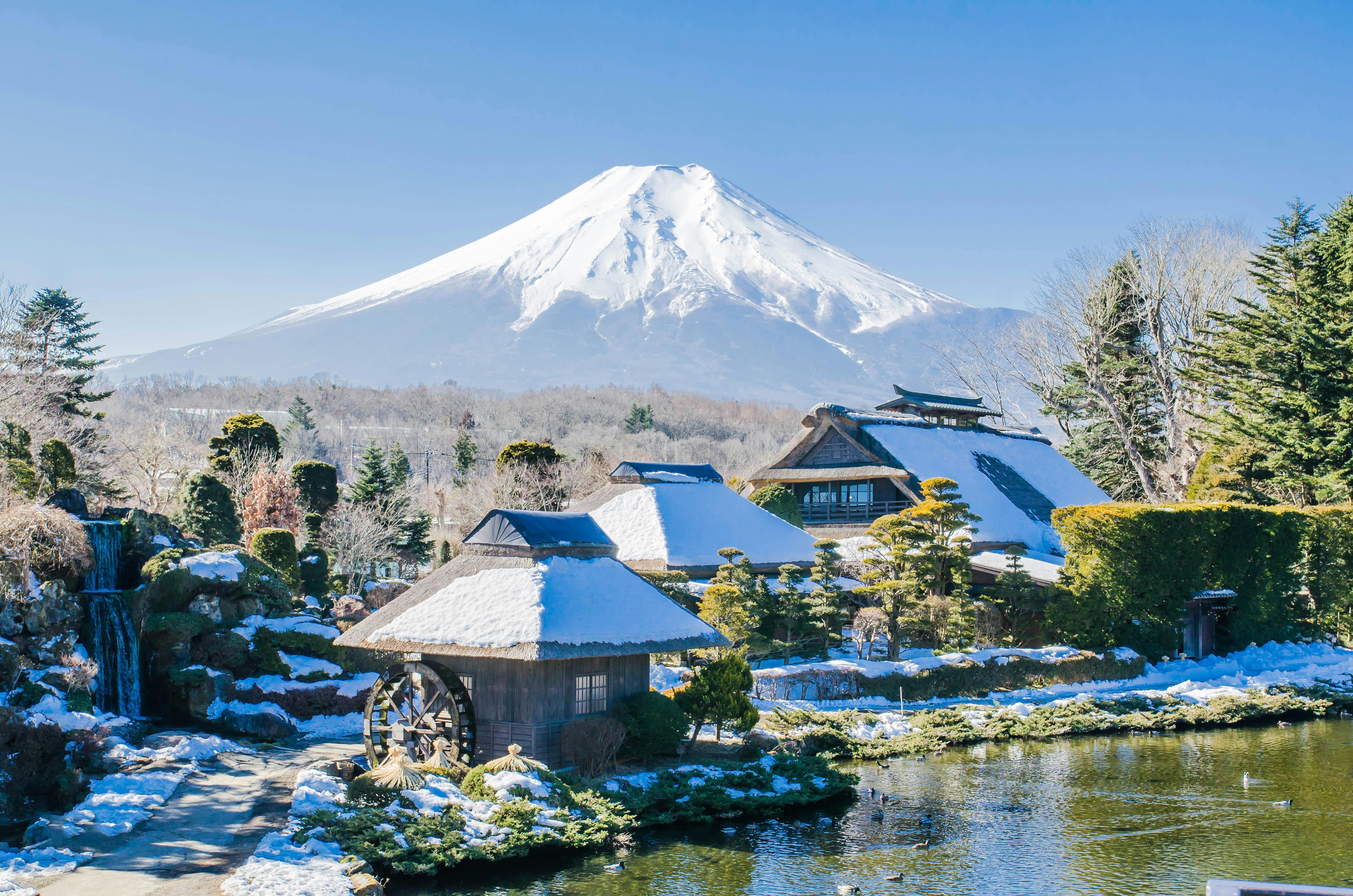 Traditional Japanese houses with thatched roofs beside a pond and a water wheel, surrounded by trees and light snow, with Mount Fuji covered in snow in the background under a clear blue sky.