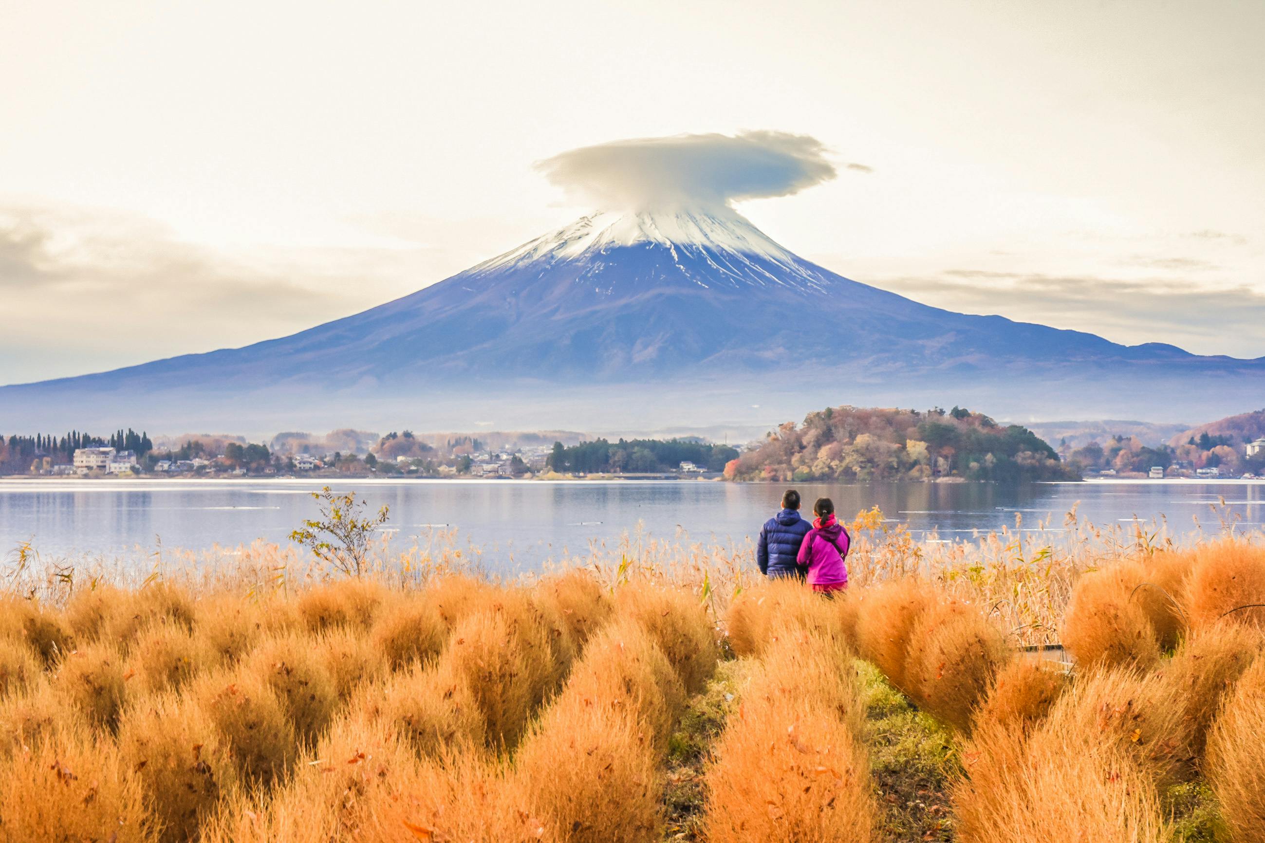 Two people stand in a field of golden grass, looking at Mount Fuji across a lake. The mountain is snow-capped with a cloud hovering over its peak, and autumn colors surround the scene.