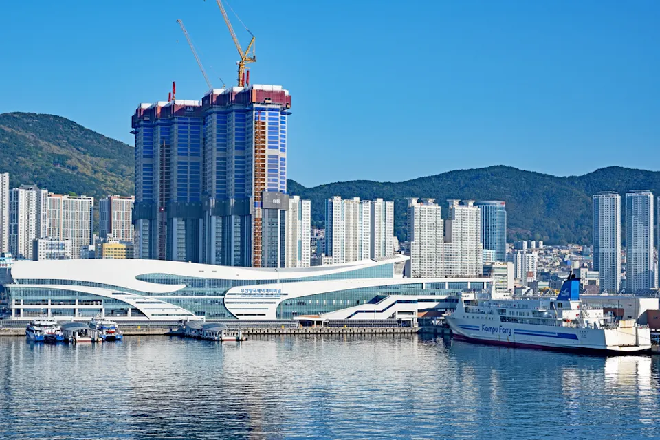 View of a coastal city skyline with a modern ferry terminal and a docked ferry labeled "Panstar Ferry." High-rise buildings and a construction site with cranes are visible against a backdrop of green hills and a clear blue sky.