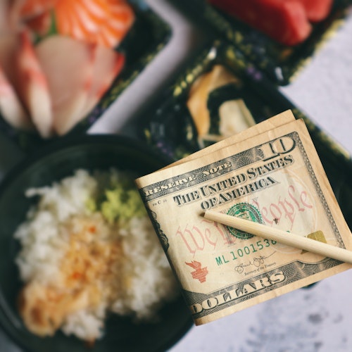 Tipping in Japan Chopsticks holding a folded 20-dollar bill over a table with assorted sushi and a bowl of rice topped with wasabi and soy sauce. Plates contain pieces of nigiri sushi with fish and shrimp.