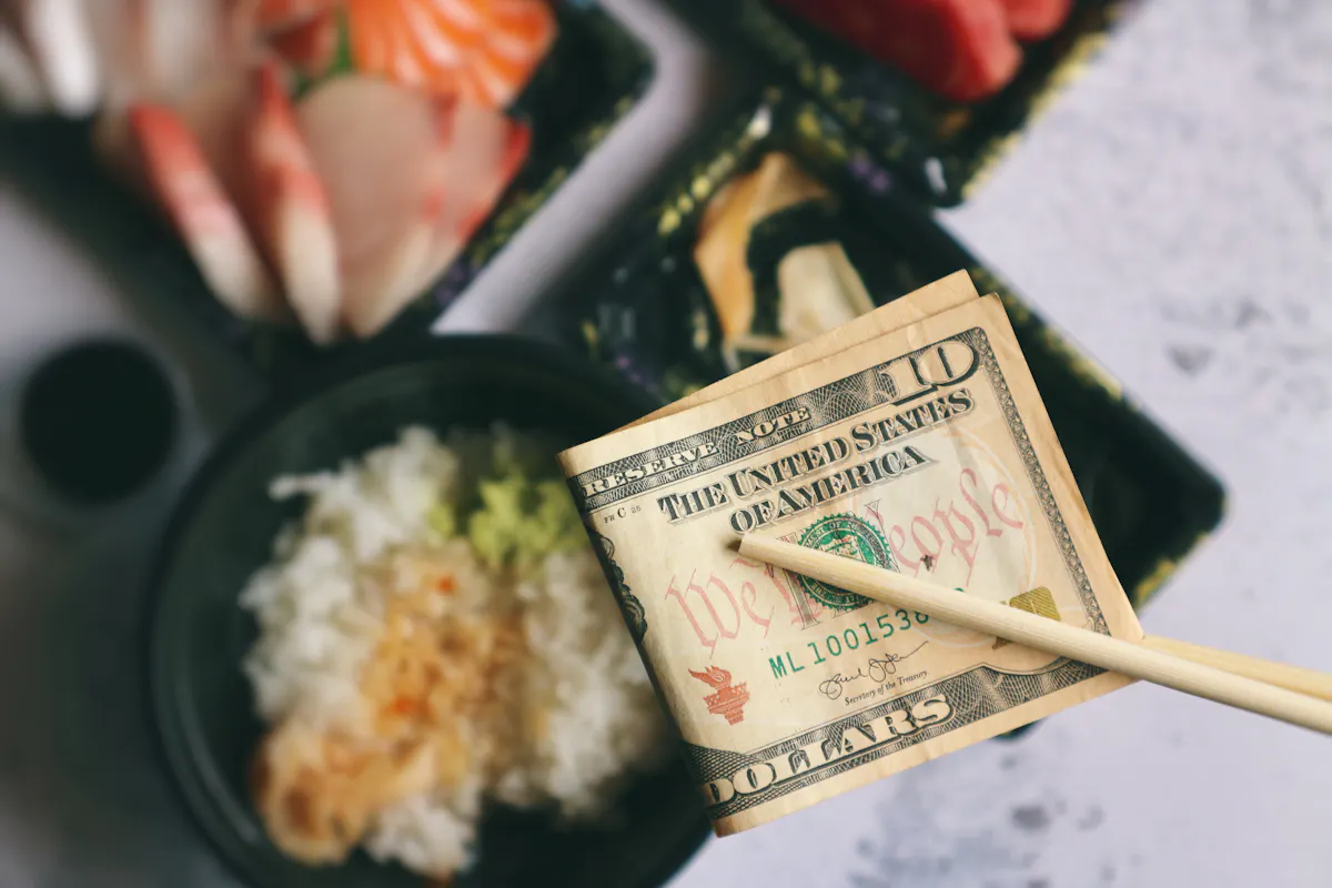 Tipping in Japan Chopsticks holding a folded 20-dollar bill over a table with assorted sushi and a bowl of rice topped with wasabi and soy sauce. Plates contain pieces of nigiri sushi with fish and shrimp.