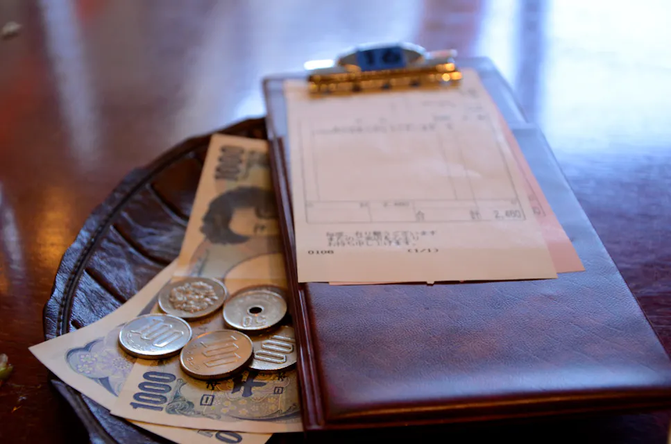 Tipping Etiquette A small tray holds Japanese yen bills and coins alongside a receipt clipped to a brown folder. The tray is placed on a wooden table, and the currency includes 1000 yen bills and various coins.