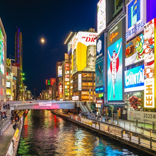 Dotonbori Osaka A vibrant cityscape of Dotonbori, Osaka, at night. Bright neon signs and billboards illuminate the bustling canal and crowded walkways. The iconic Glico Running Man sign is prominently featured, reflecting in the water below.