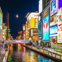 Dotonbori Osaka A vibrant cityscape of Dotonbori, Osaka, at night. Bright neon signs and billboards illuminate the bustling canal and crowded walkways. The iconic Glico Running Man sign is prominently featured, reflecting in the water below.