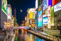 A vibrant cityscape of Dotonbori, Osaka, at night. Bright neon signs and billboards illuminate the bustling canal and crowded walkways. The iconic Glico Running Man sign is prominently featured, reflecting in the water below.