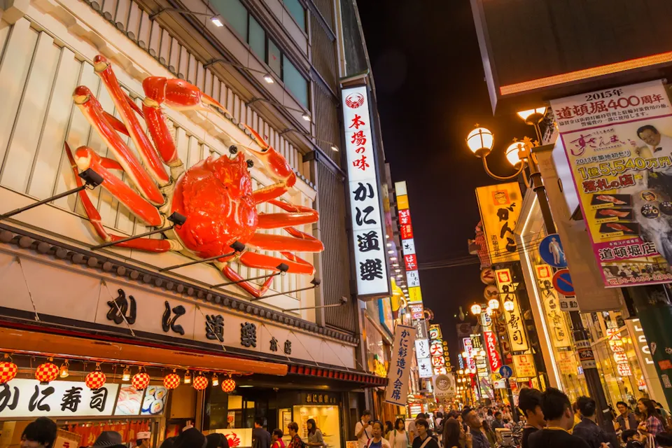 Bustling street in Dotonbori, Osaka, at night. A large red crab sign adorns a building, with bright lights illuminating various shop signs. Crowds of people walk along the lively street, enjoying the vibrant atmosphere.