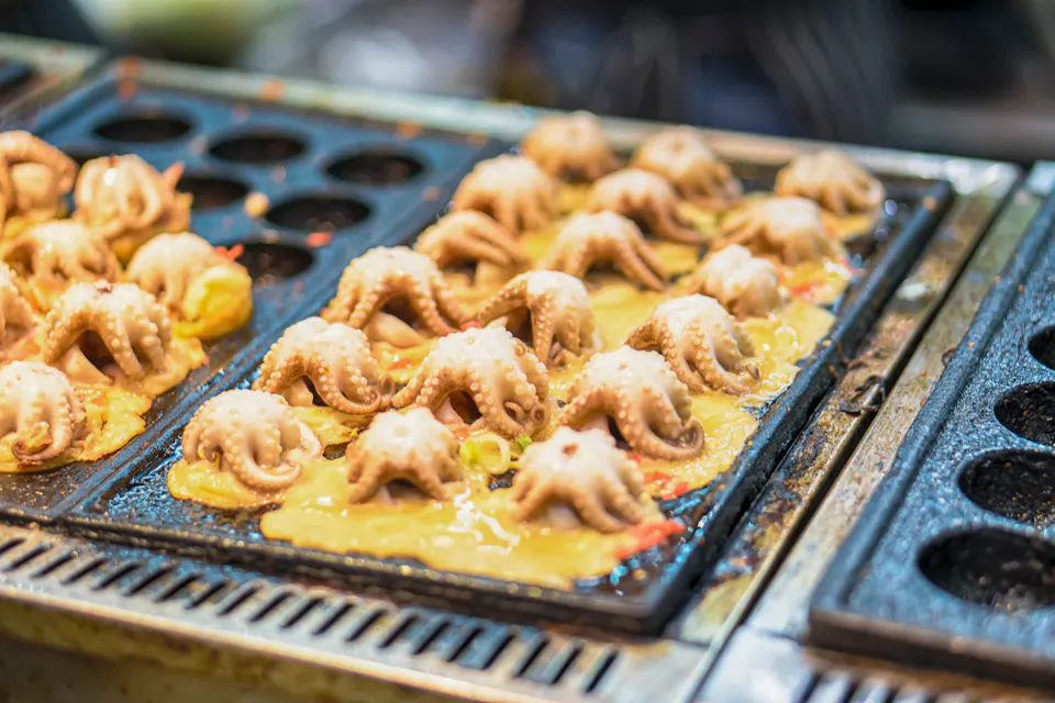 Close-up of octopus-shaped snacks cooking in a takoyaki pan. The dough is bubbling around each piece, and the pan is set on a grill. The background shows a slightly blurred kitchen setting.