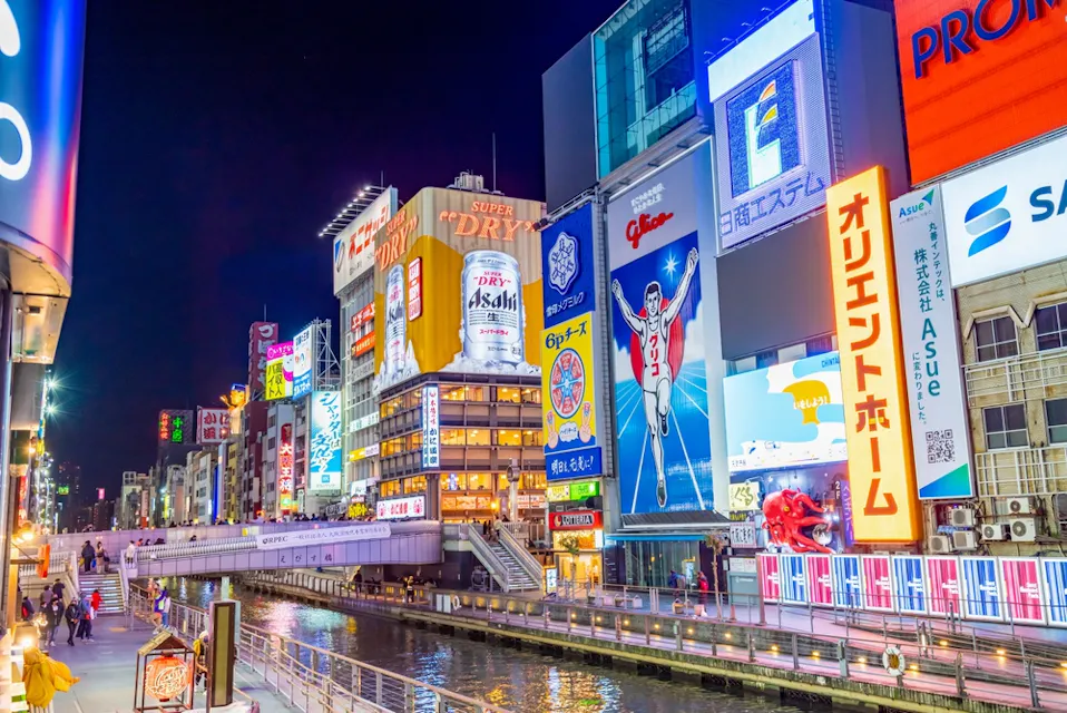 A vibrant nighttime scene in Dotonbori, Osaka, Japan, featuring illuminated billboards and neon signs lining a canal. Pedestrians walk along the waterway under the bright advertising, creating a lively urban atmosphere.