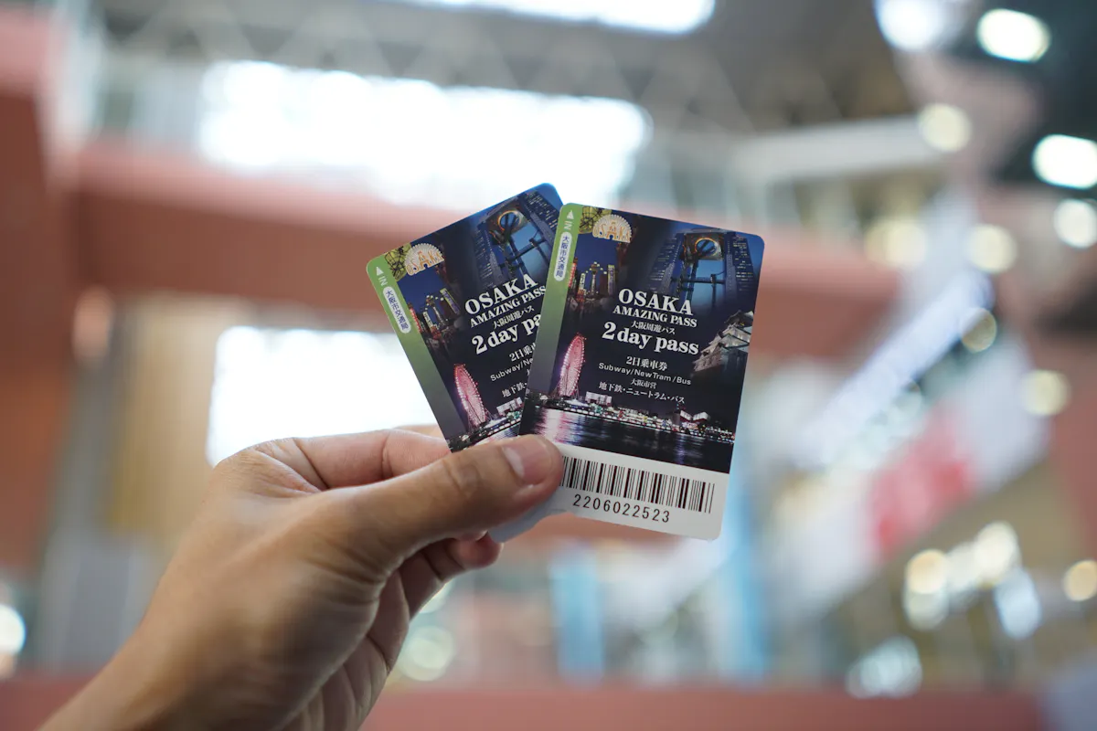 Osaka Amazing Pass A hand holds two Osaka Amazing Pass 2-day tickets against a blurred background of what appears to be an indoor setting. The tickets feature cityscape imagery with a ferris wheel and other attractions.
