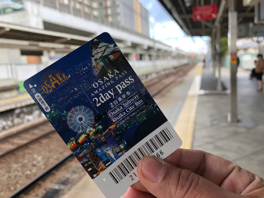 What Is the Osaka Amazing Pass A person holds an "Osaka Amazing Pass" for 2 days, valid for Osaka Subway and City Bus, at a train station platform. The pass features images of Osaka attractions and a barcode at the bottom. The railway track extends into the distance.