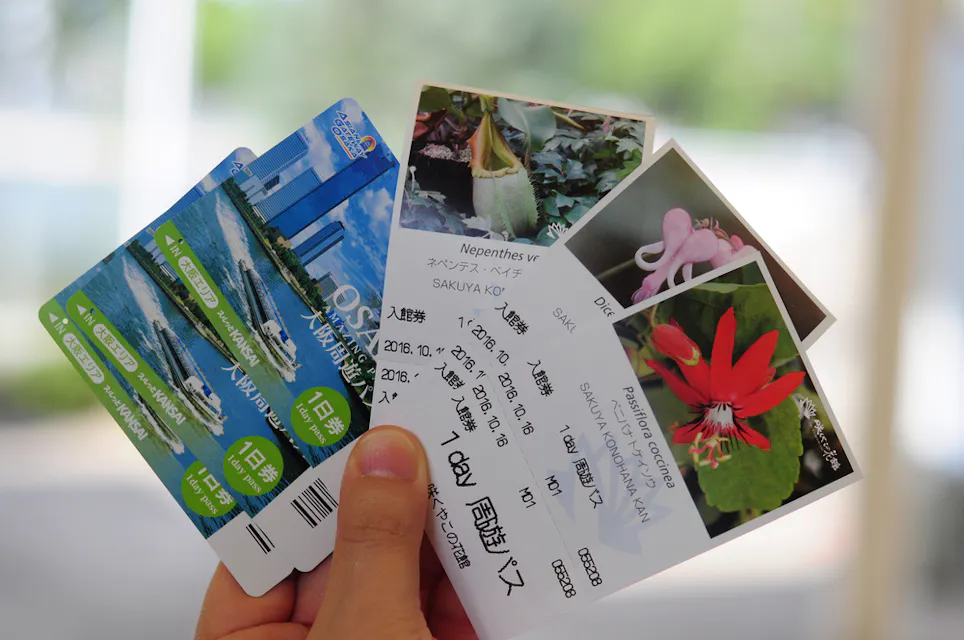 Benefits of the Osaka Amazing Pass A hand holding four colorful tickets, each displaying different scenic images and text. The tickets have Asian characters and depict a variety of attractions, including plants and a cityscape with water and buildings in the background.