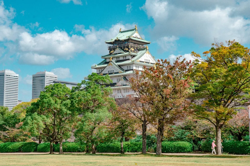 Suggested Itinerary A picturesque scene of a traditional Japanese castle with green and golden roof tiles, surrounded by lush trees under a vibrant blue sky. Modern skyscrapers peek in the background, blending historic and contemporary architecture.