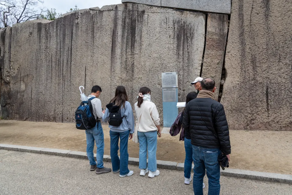 A small group of people are gathered in front of a large stone wall. They appear to be reading or observing a plaque affixed to the wall. Some are wearing jackets and carrying backpacks. The scene is set outdoors on a paved pathway.