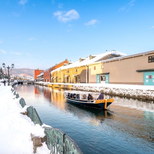 Visiting Japan in January A boat with people is floating along a snow-covered canal, flanked by snowy sidewalks and historic brick buildings under a clear blue sky. A few people are walking on the left path, and trees are visible in the distance.