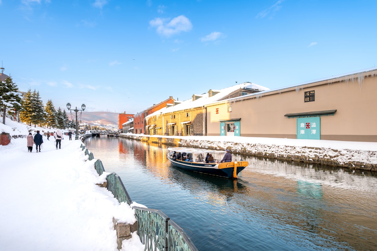 Visiting Japan in January A boat with people is floating along a snow-covered canal, flanked by snowy sidewalks and historic brick buildings under a clear blue sky. A few people are walking on the left path, and trees are visible in the distance.