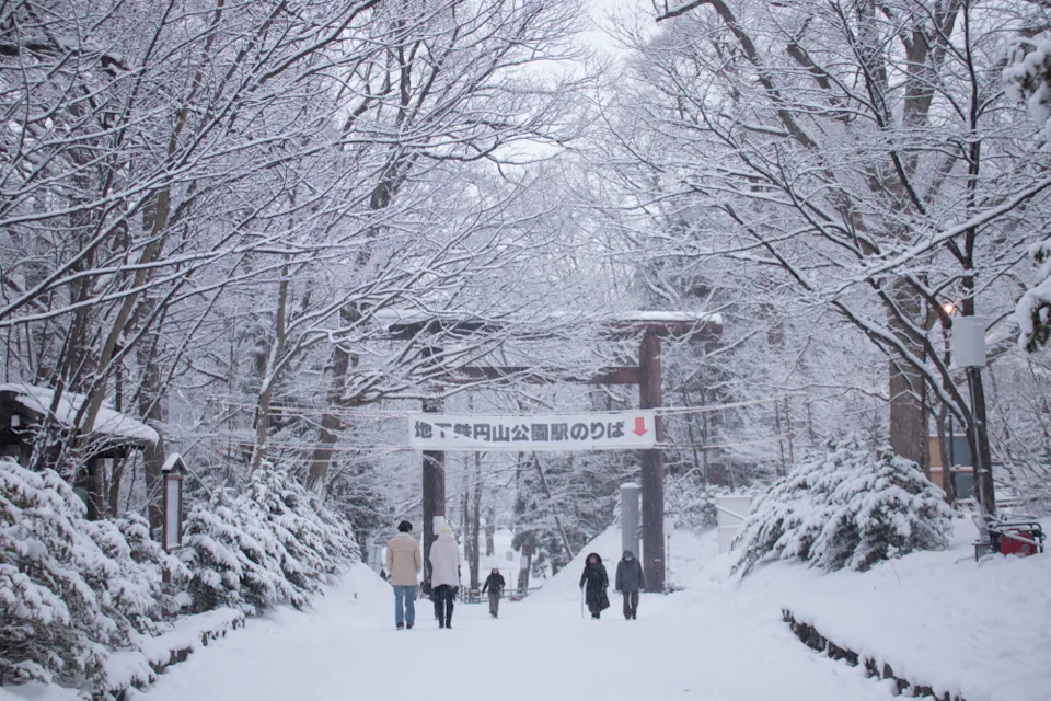 Weather in Japan During January A snowy path in a park, lined with bare trees covered in snow. A group of people walks towards a wooden torii gate with a sign above it. Snow blankets the ground and bushes, creating a serene winter scene.