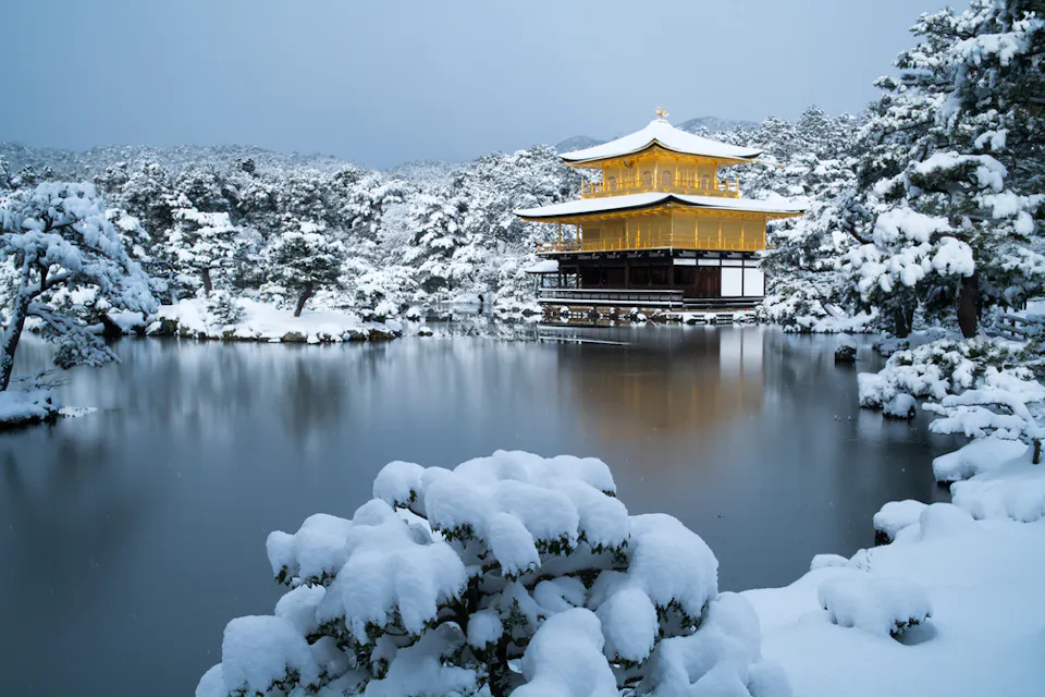 Winter Season in Japan A serene winter scene of Kinkaku-ji, a golden pavilion, reflected in a tranquil pond surrounded by snow-laden trees. The sky is overcast, adding to the peaceful atmosphere of the snowy landscape.