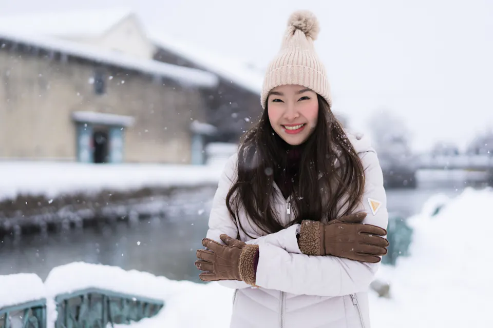 What to Pack for January in Japan A woman wearing a beige knit hat and brown gloves stands smiling with her arms crossed in a snowy outdoor setting. She is dressed in a light-colored winter coat, and snow gently falls around her near a blurred building and waterway.