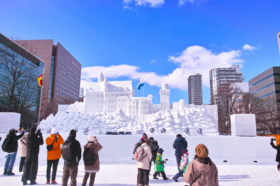 Things to Do in Japan During January A group of people stands in front of a large, intricate snow sculpture depicting a castle or palace. The structure is surrounded by smaller snow formations. Tall buildings and a clear blue sky with scattered clouds serve as the backdrop.