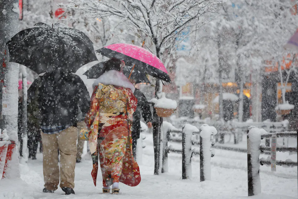 A person in a colorful kimono walks with another person under umbrellas on a snowy street. Snow covers the trees and sidewalks, creating a wintry urban scene.