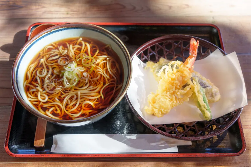A bowl of noodle soup garnished with green onions sits on a tray beside a plate of assorted tempura, including a prawn and vegetables. Sunlight streams onto the wooden table, highlighting the meal.