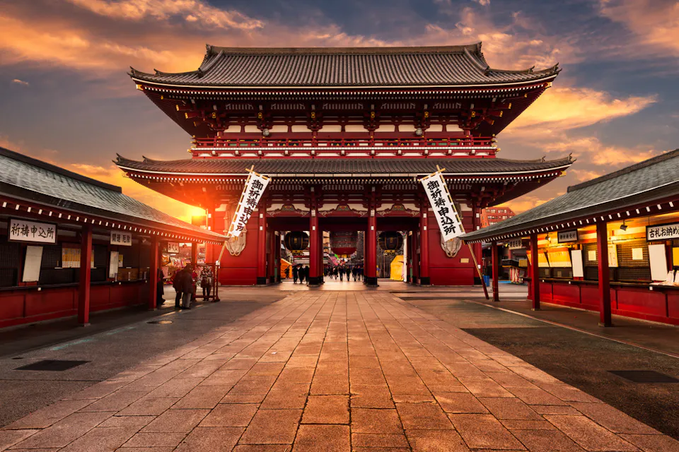 The image shows a view of Senso-ji Temple in Tokyo at sunset. The temple's red and white entrance is prominently featured, with a paved walkway leading up to it. The sky is vibrant with hues of orange and yellow.