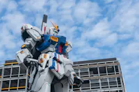A large Gundam statue stands prominently against a backdrop of a partly cloudy sky, with a building partially visible behind it. The robot features blue, white, and red colors with a yellow accent on its head.