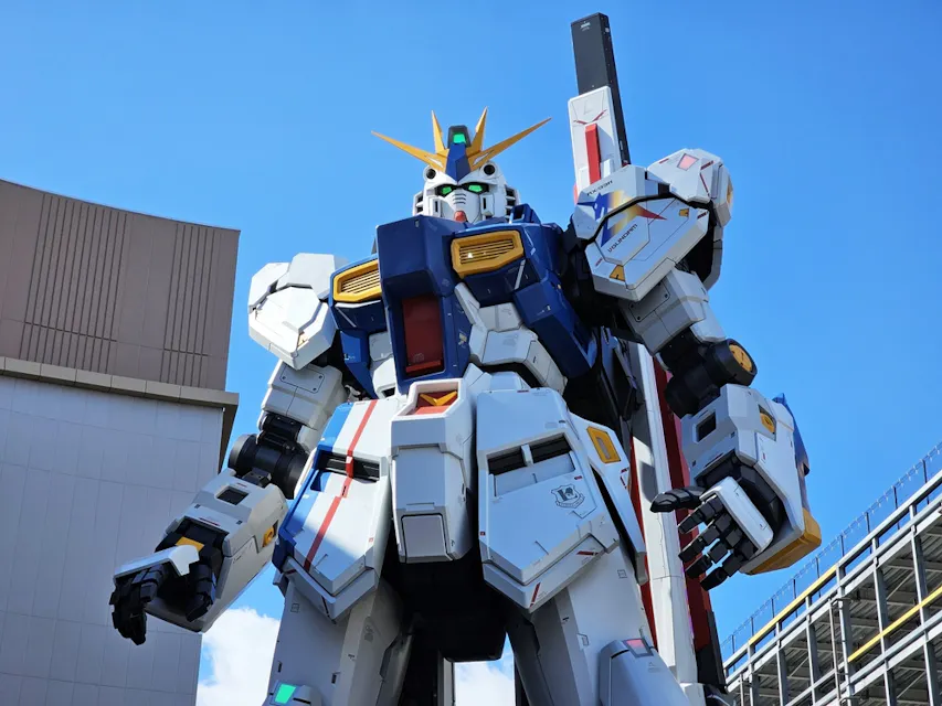 A large, white, blue, and yellow humanoid robot statue stands against a clear blue sky. It features intricate details and mechanical elements. Buildings are visible in the background.
