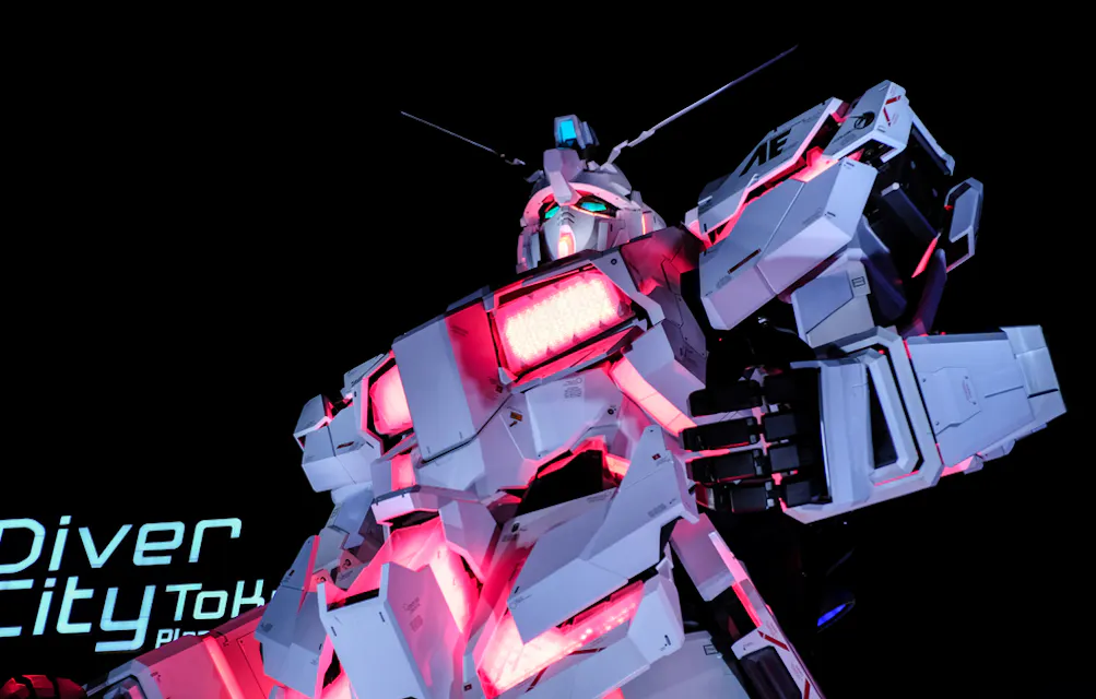 A large, illuminated robot statue with glowing red and white lights stands against a dark sky. Its intricate design showcases a futuristic and mechanical appearance. A partial sign reads "Diver City Tokyo Plaza" in the background.