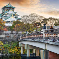 Osaka Castle A scenic view of Osaka Castle in Japan during autumn. The historic castle towers above colorful foliage and trees. A traditional wooden bridge crosses a tranquil moat in the foreground under a partly cloudy sky.