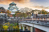 A scenic view of Osaka Castle in Japan during autumn. The historic castle towers above colorful foliage and trees. A traditional wooden bridge crosses a tranquil moat in the foreground under a partly cloudy sky.
