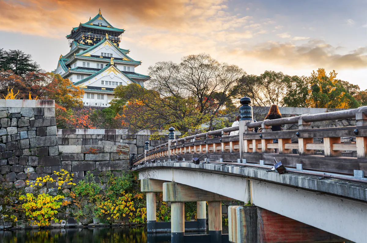 A scenic view of Osaka Castle in Japan during autumn. The historic castle towers above colorful foliage and trees. A traditional wooden bridge crosses a tranquil moat in the foreground under a partly cloudy sky.