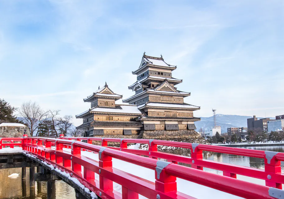 A traditional Japanese castle with multiple tiered rooftops stands beside a calm water moat. A vibrant red bridge leads toward the castle. Snow covers parts of the bridge and surrounding landscape, with mountains in the background under a clear sky.