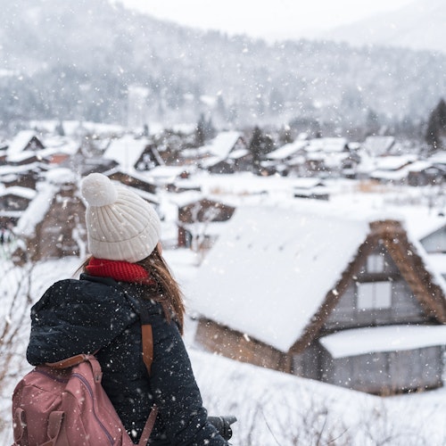 Snow in Japan A person with a pink backpack and a white beanie looks at a snowy village with traditional houses. Snowflakes fall gently, creating a serene winter scene. Trees and mountains are visible in the background.