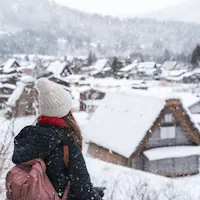 Snow in Japan A person with a pink backpack and a white beanie looks at a snowy village with traditional houses. Snowflakes fall gently, creating a serene winter scene. Trees and mountains are visible in the background.