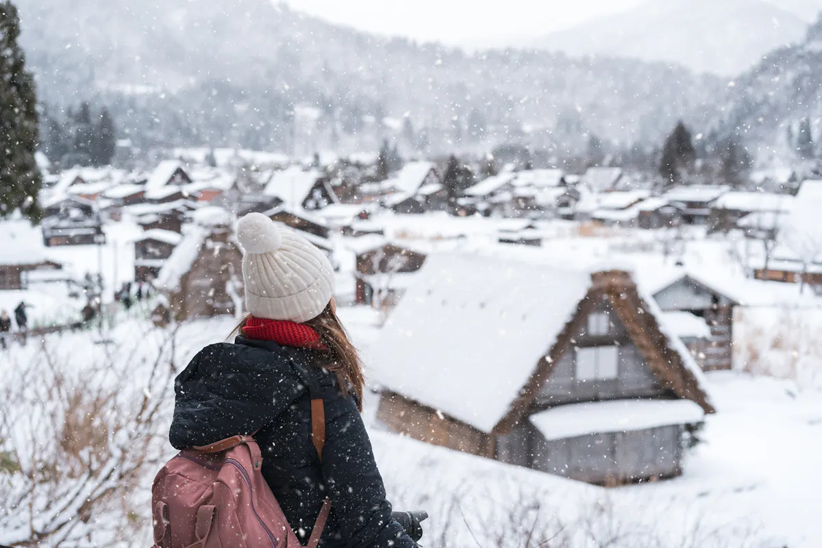 Snow in Japan A person with a pink backpack and a white beanie looks at a snowy village with traditional houses. Snowflakes fall gently, creating a serene winter scene. Trees and mountains are visible in the background.