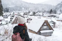 A person with a pink backpack and a white beanie looks at a snowy village with traditional houses. Snowflakes fall gently, creating a serene winter scene. Trees and mountains are visible in the background.