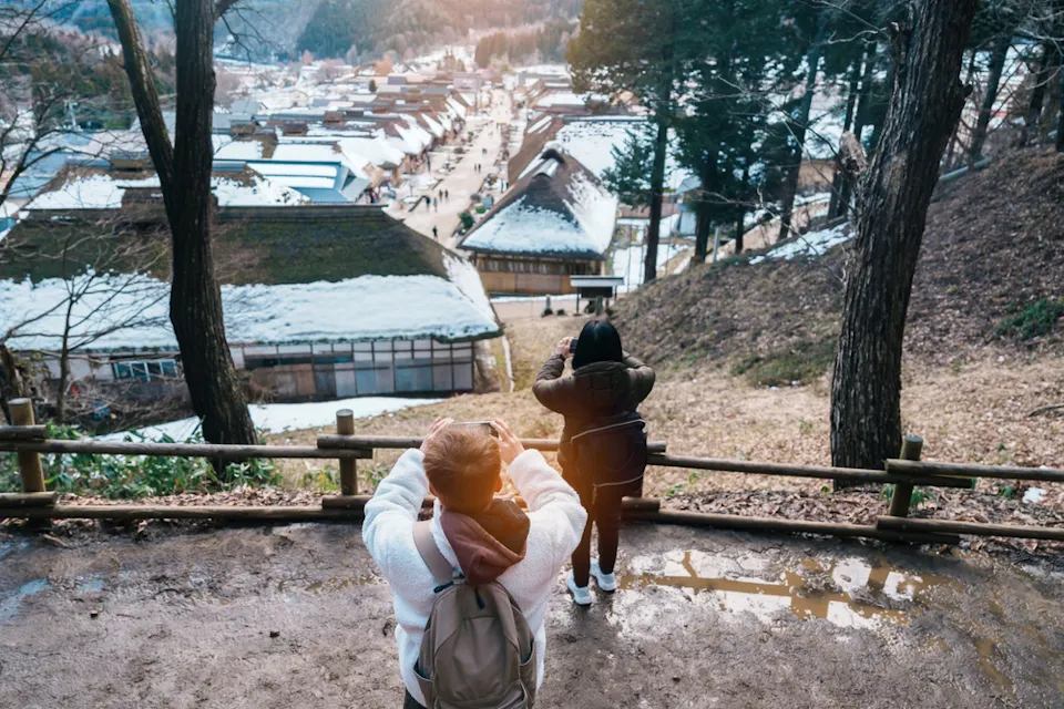Two people stand on a hilltop, photographing a snow-covered village surrounded by trees. The village features traditional wooden buildings with thatched roofs, and the scene is set in a tranquil, wintry landscape.