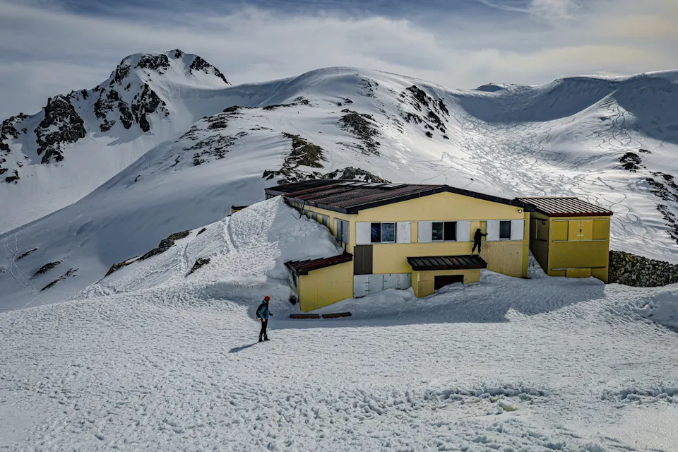 A person walks toward a yellow mountain hut partially covered in snow, located on a snowy mountain slope. Rugged snow-covered peaks and cloudy skies are visible in the background.