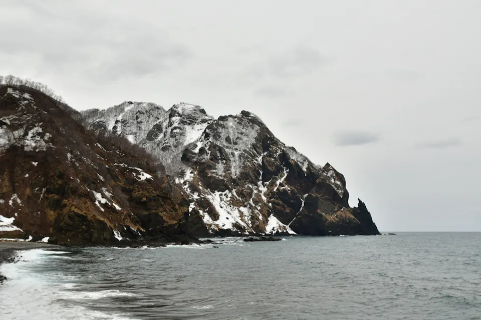 A rugged, rocky coastline with patches of snow overlooking a calm, grey sea under an overcast sky. The terrain is steep, and the water is slightly rippled near the shore. Sparse vegetation is visible on the cliffs.