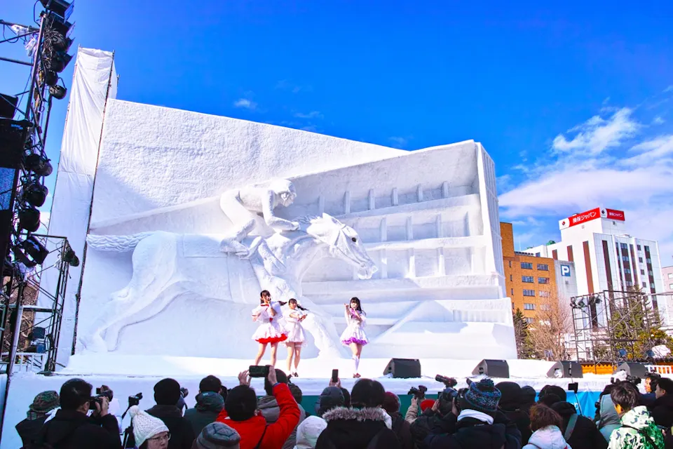 A large snow sculpture of a galloping horse and rider is displayed on an outdoor stage. Three performers in colorful outfits sing and dance in front of it, while an audience photographs the scene. Tall buildings and a clear blue sky are in the background.