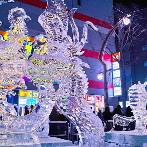 Japan in February Intricate ice sculptures of mythical creatures, including a phoenix and dragon, glow with colorful lights in an urban setting. A crowd observes the artwork against a background of illuminated buildings and signs at night.