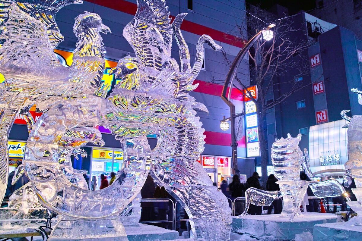 Japan in February Intricate ice sculptures of mythical creatures, including a phoenix and dragon, glow with colorful lights in an urban setting. A crowd observes the artwork against a background of illuminated buildings and signs at night.