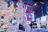 Intricate ice sculptures of mythical creatures, including a phoenix and dragon, glow with colorful lights in an urban setting. A crowd observes the artwork against a background of illuminated buildings and signs at night.