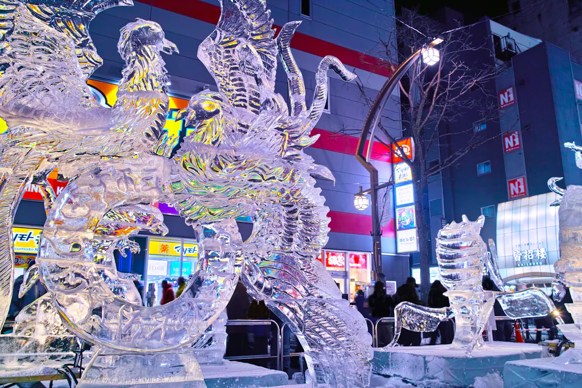 Intricate ice sculptures of mythical creatures, including a phoenix and dragon, glow with colorful lights in an urban setting. A crowd observes the artwork against a background of illuminated buildings and signs at night.