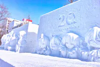 Snow sculptures depicting various anime characters are showcased at a winter festival. One sculpture features "20th Anniversary" engraved on it. Snow covers the ground and trees, while buildings are visible in the background under a clear blue sky.