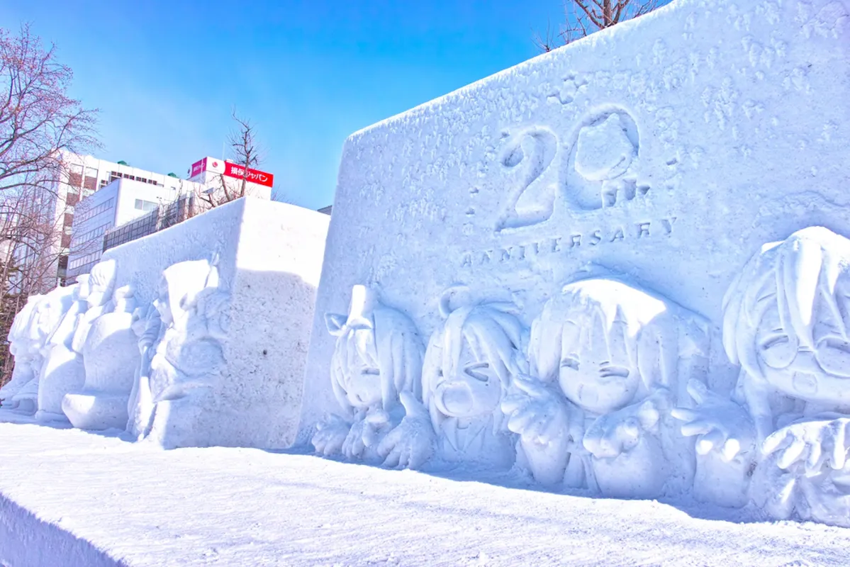 Snow sculptures depicting various anime characters are showcased at a winter festival. One sculpture features "20th Anniversary" engraved on it. Snow covers the ground and trees, while buildings are visible in the background under a clear blue sky.