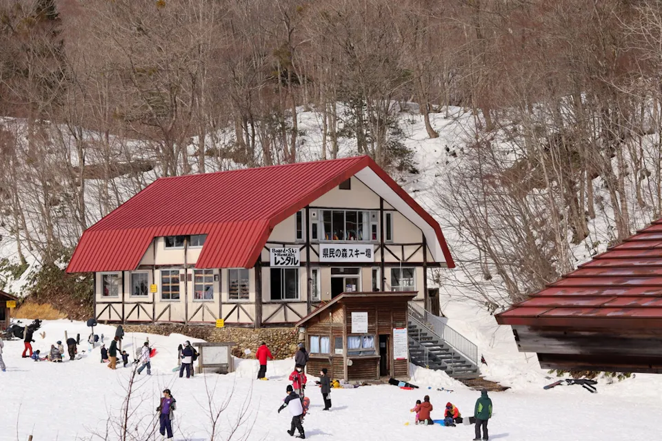 A snow-covered ski resort with people enjoying winter activities. The main building features a red roof and wood accents. Visitors are skiing, snowboarding, and playing in the snow, surrounded by leafless trees and a forested hill in the background.