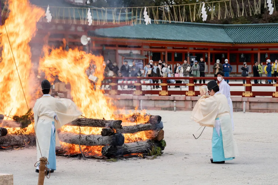 People in traditional clothing participate in a fire ceremony, standing near large burning logs. A crowd watches from behind a barrier. The event takes place in an open area with traditional architecture in the background.