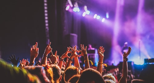 Audience at a concert with colorful lights in the background. People raise their hands, some forming a heart shape. The atmosphere is energetic and lively.